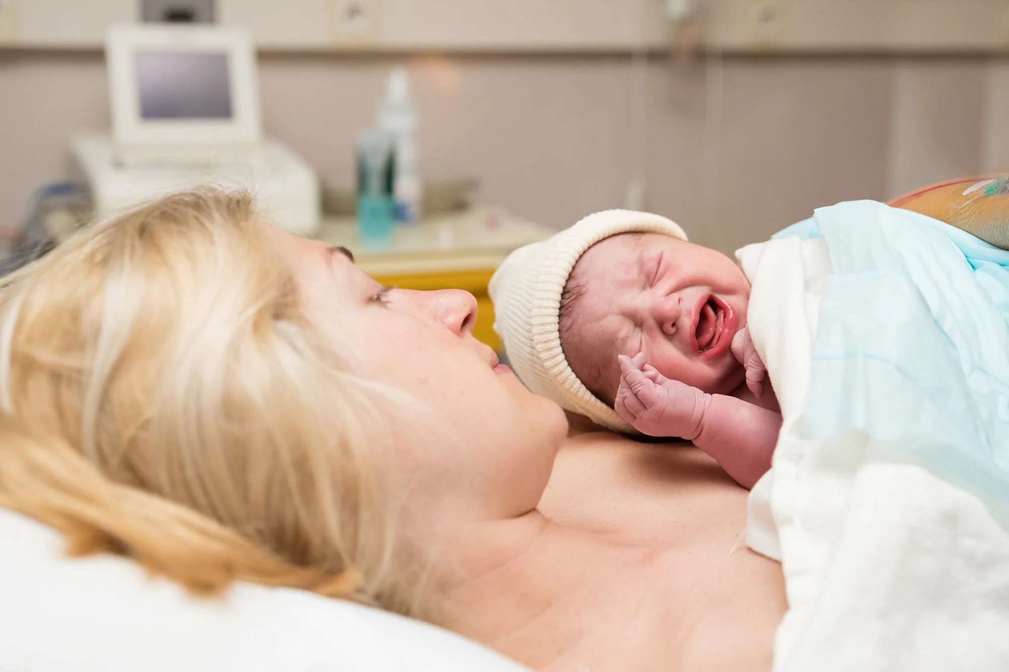 A newborn baby crying while lying on a woman's chest in a hospital room.