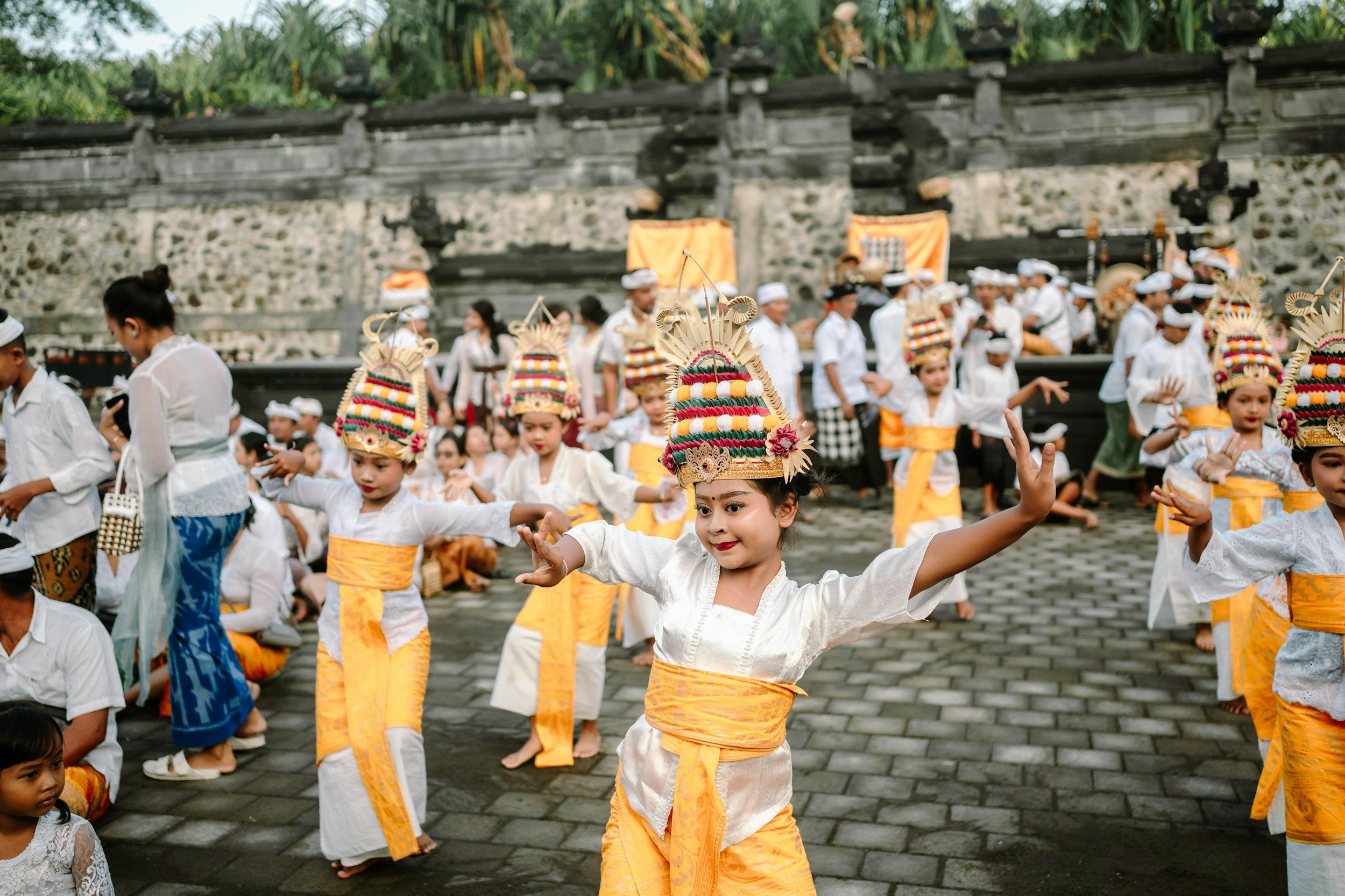 Children dressed in traditional Balinese attire dancing and performing in a cultural ceremony outdoors with stone walls and greenery in the background.