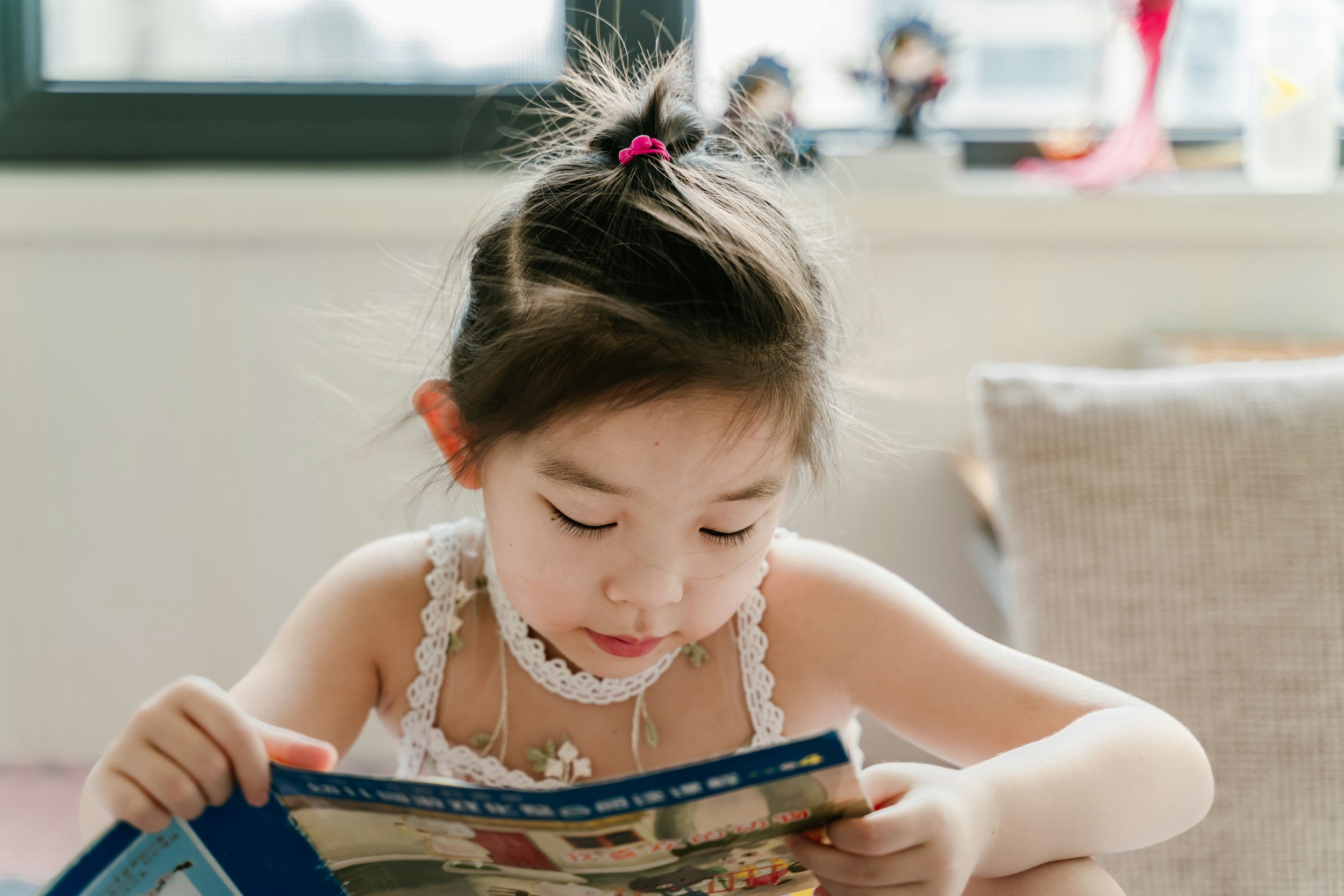 A young girl with brown hair tied with a pink scrunchie, reading a comic book or magazine at a table in a bright room.