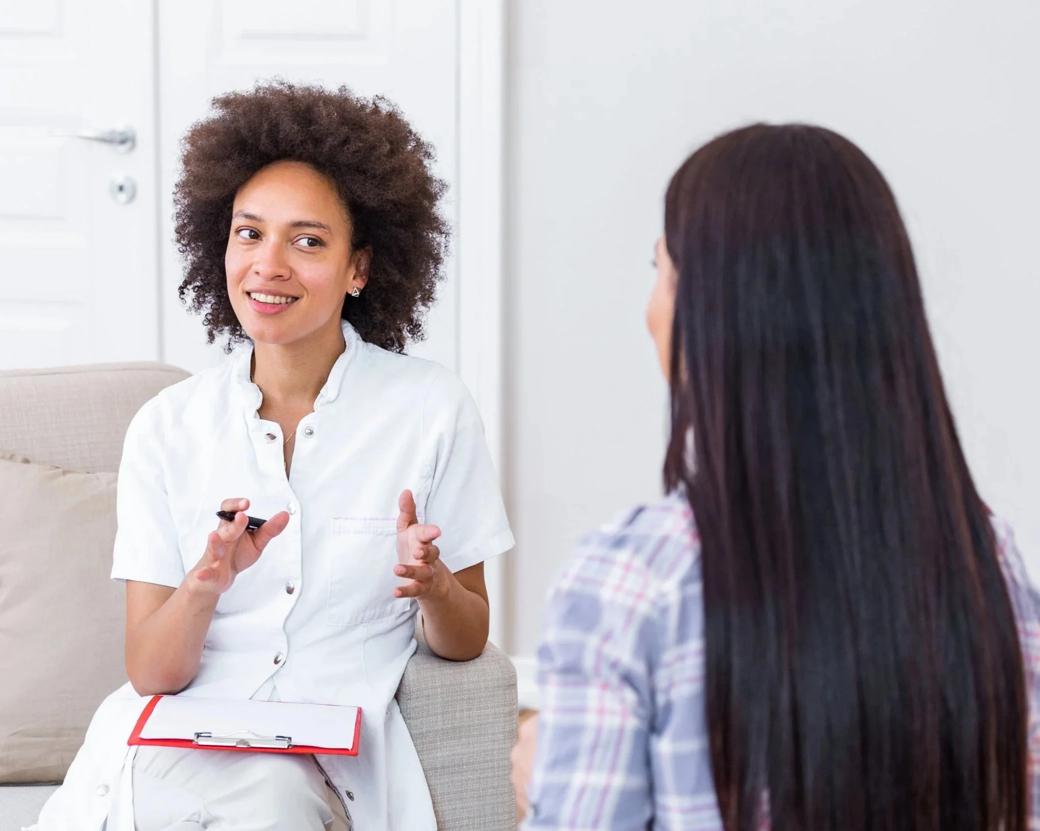 A woman in white medical scrubs holding a clipboard and gesturing with her hands, talking to another woman with long dark hair, in a bright room.