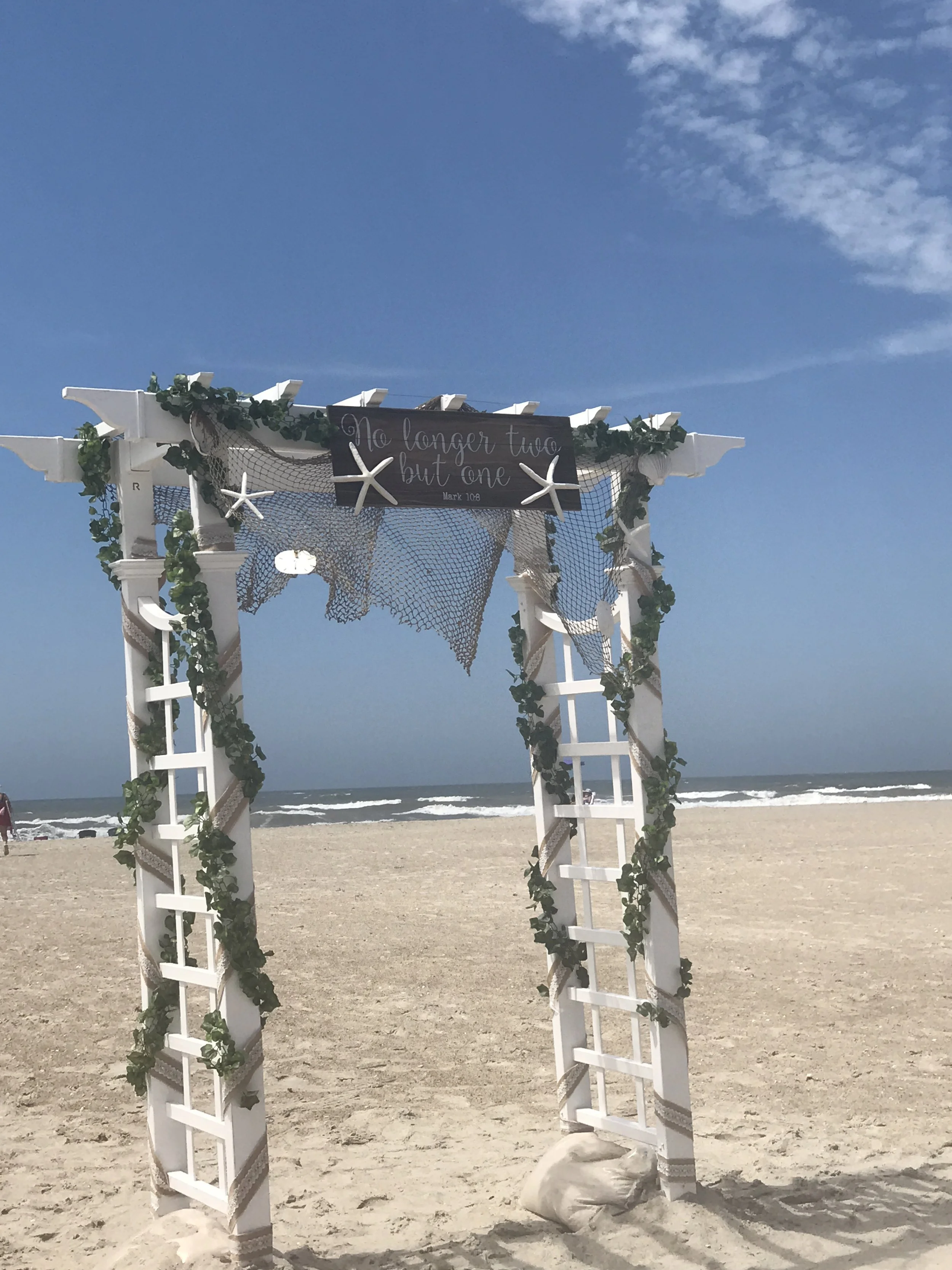 Decorated wedding arch on the beach with a sign that says 'No longer two, but one,' featuring starfish, greenery, and netting, under a blue sky.