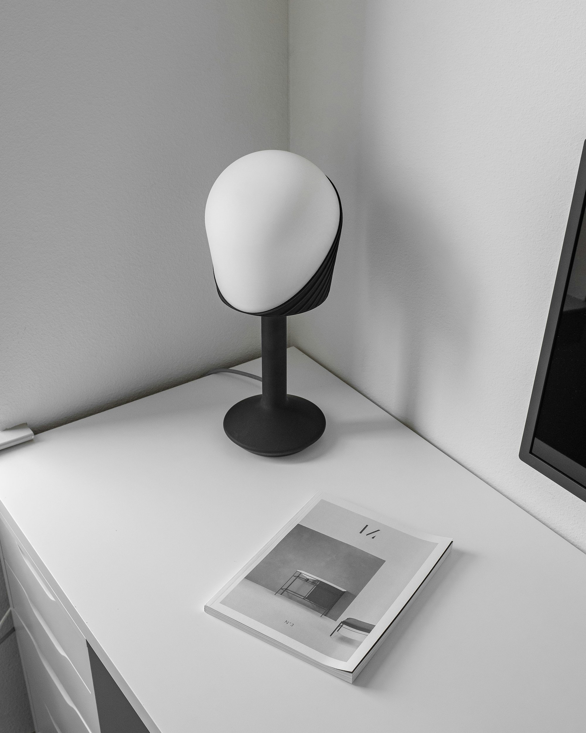 A modern black and white desk lamp on a white desk with a magazine and a part of a computer monitor.
