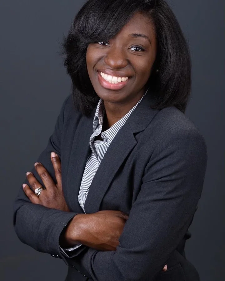 Businesswoman with black hair smiling, wearing a gray blazer and a striped shirt, arms crossed against a dark background.
