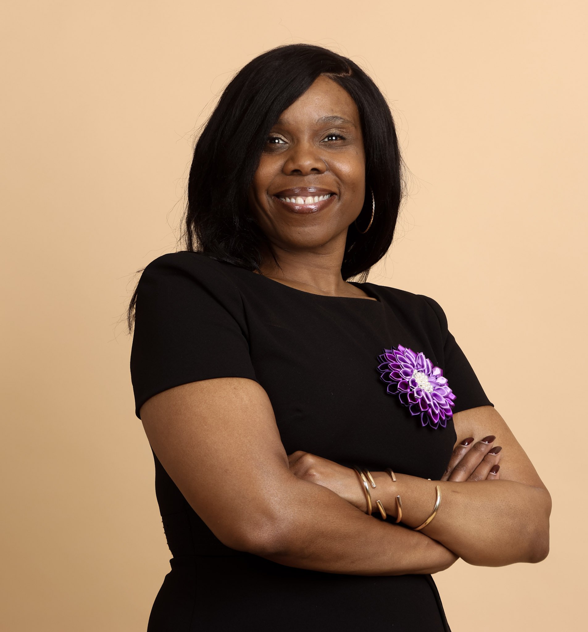A woman with black hair, wearing a black dress with a purple flower brooch, standing with arms crossed and smiling against a beige background.