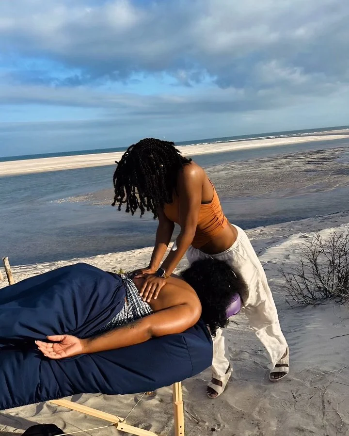 A woman lying face down on a massage table on the beach while another woman performs a massage. The setting is a sandy beach with ocean waves and a cloudy sky in the background.
