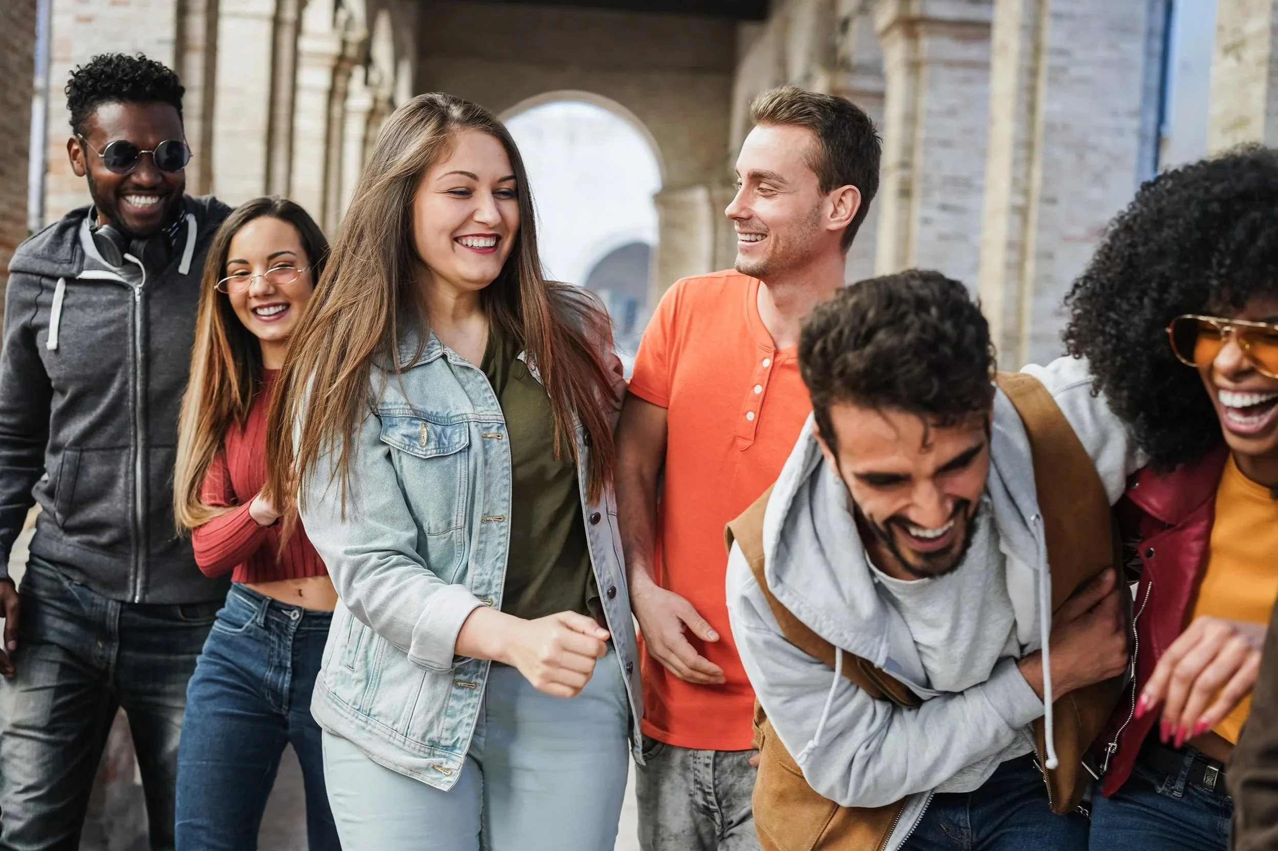 A group of seven diverse young adults smiling and laughing as they walk together outdoors in an urban area with old brick buildings.
