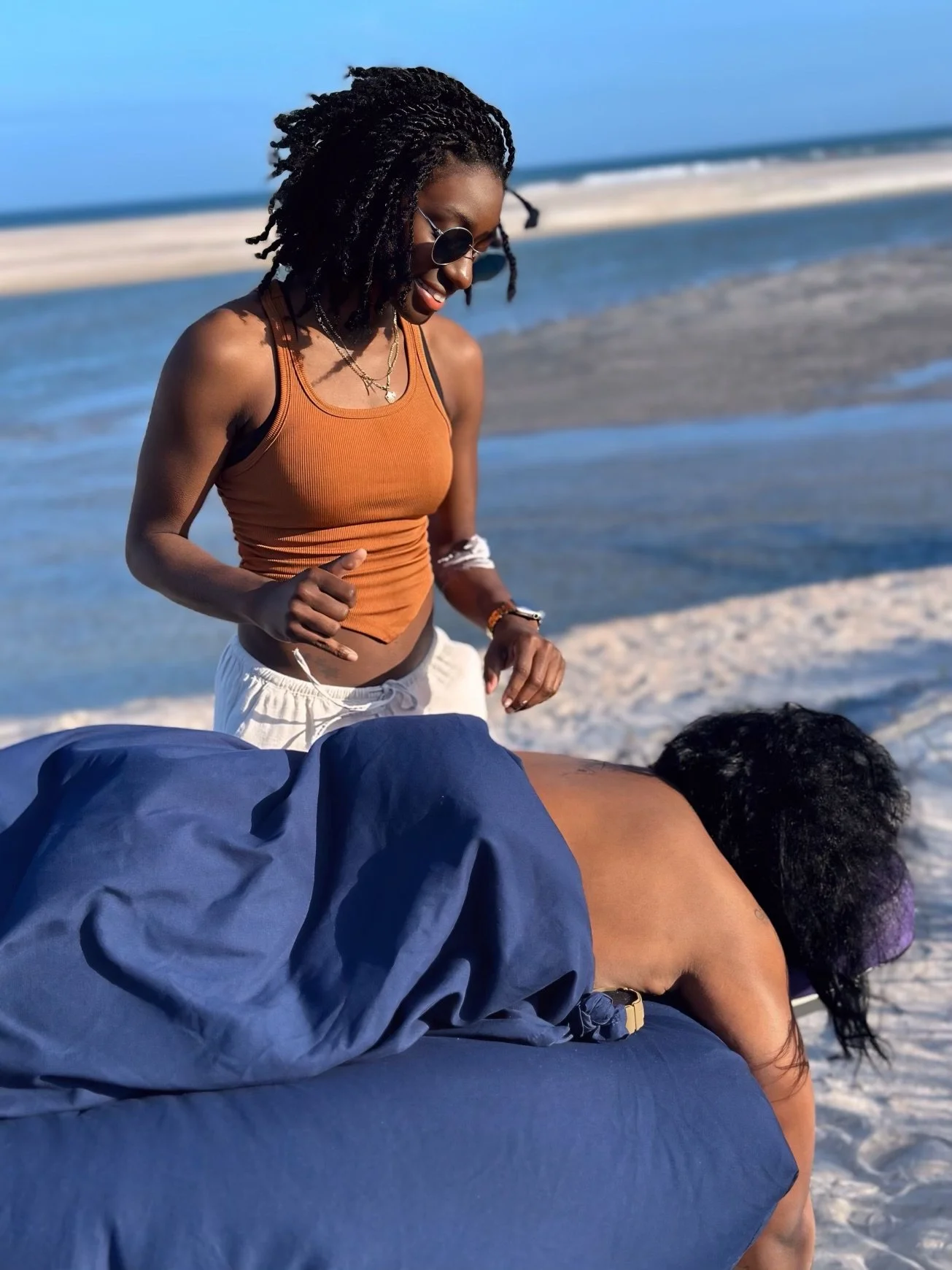 A woman in an orange tank top and sunglasses standing on a beach, looking at another person lying on a blue surface with their head turned away, with sandy and water landscape in the background.