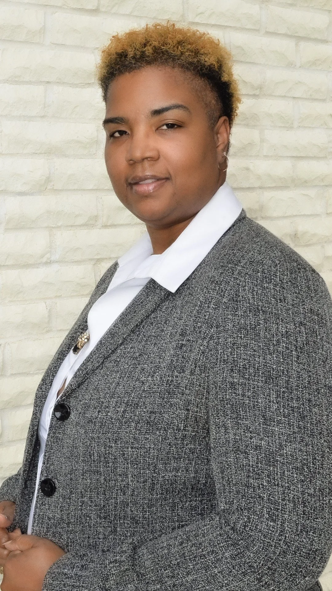 A woman with short, curly, blonde hair wearing a gray blazer over a white shirt, standing against a light-colored brick wall.