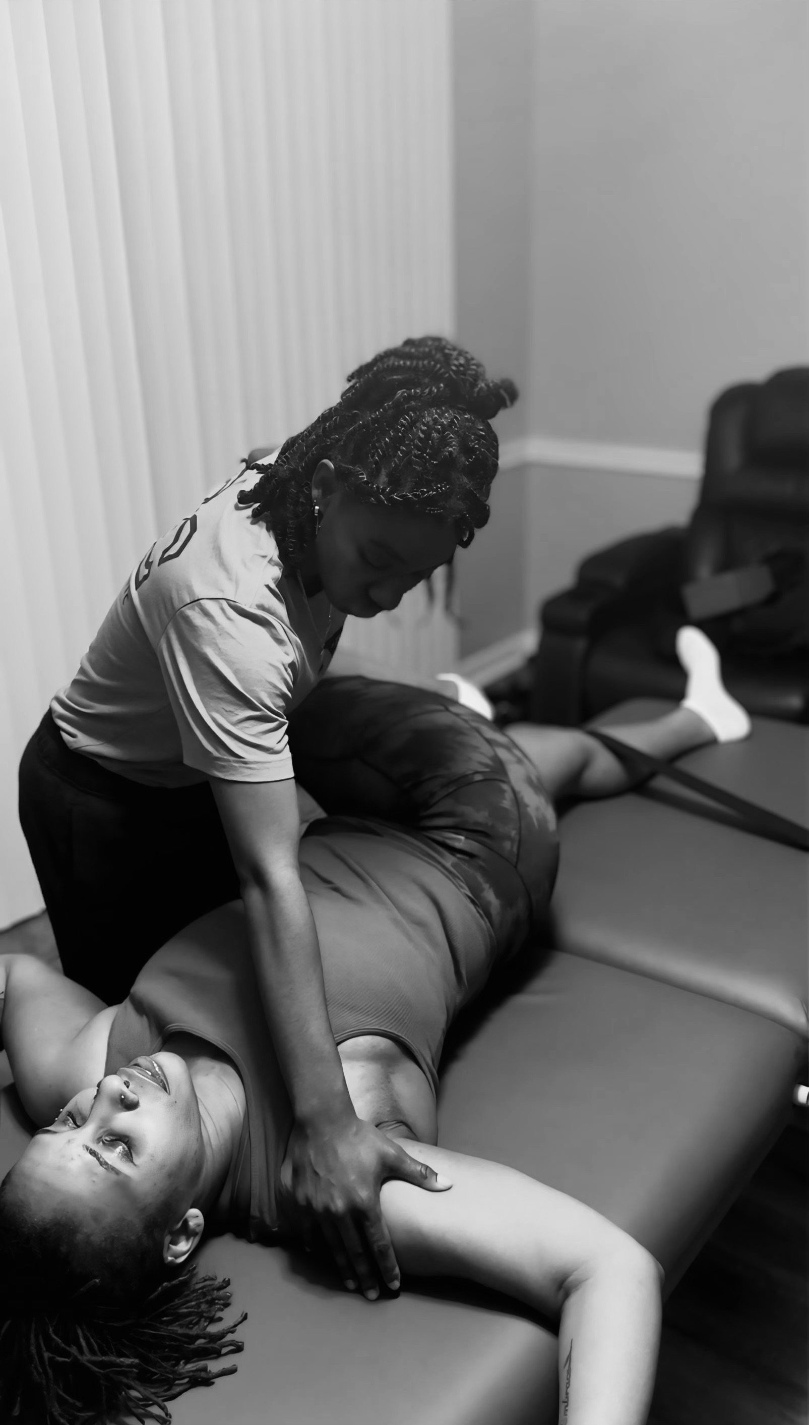 A woman receiving a massage while lying on a massage table in a room with a vertical blinds and armchair.