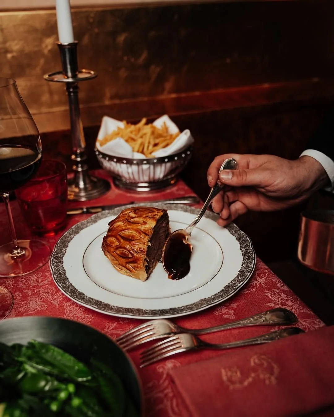 A slice of chocolate cake on a white plate with a decorative silver border, with a person's hand holding a spoon over the cake. There is a glass of red wine, a bowl of French fries, a salad, and a candle on the table, which is covered with a red patt