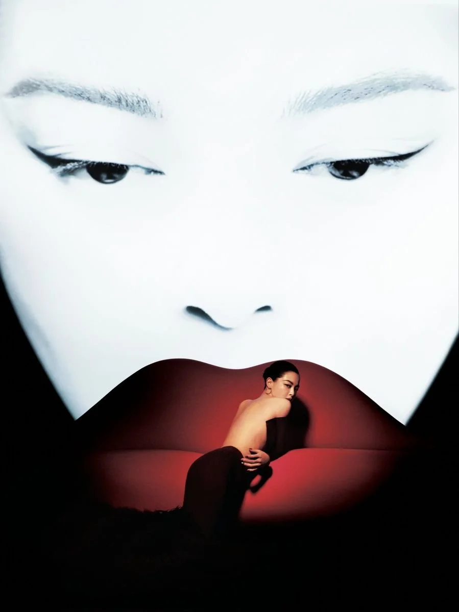 A large, high-contrast black-and-white projection of a woman's face with closed eyes, with a smaller, full-color image of a woman with dark hair and a bare back sitting on a curved, red-lit surface, positioned in front of the projection.