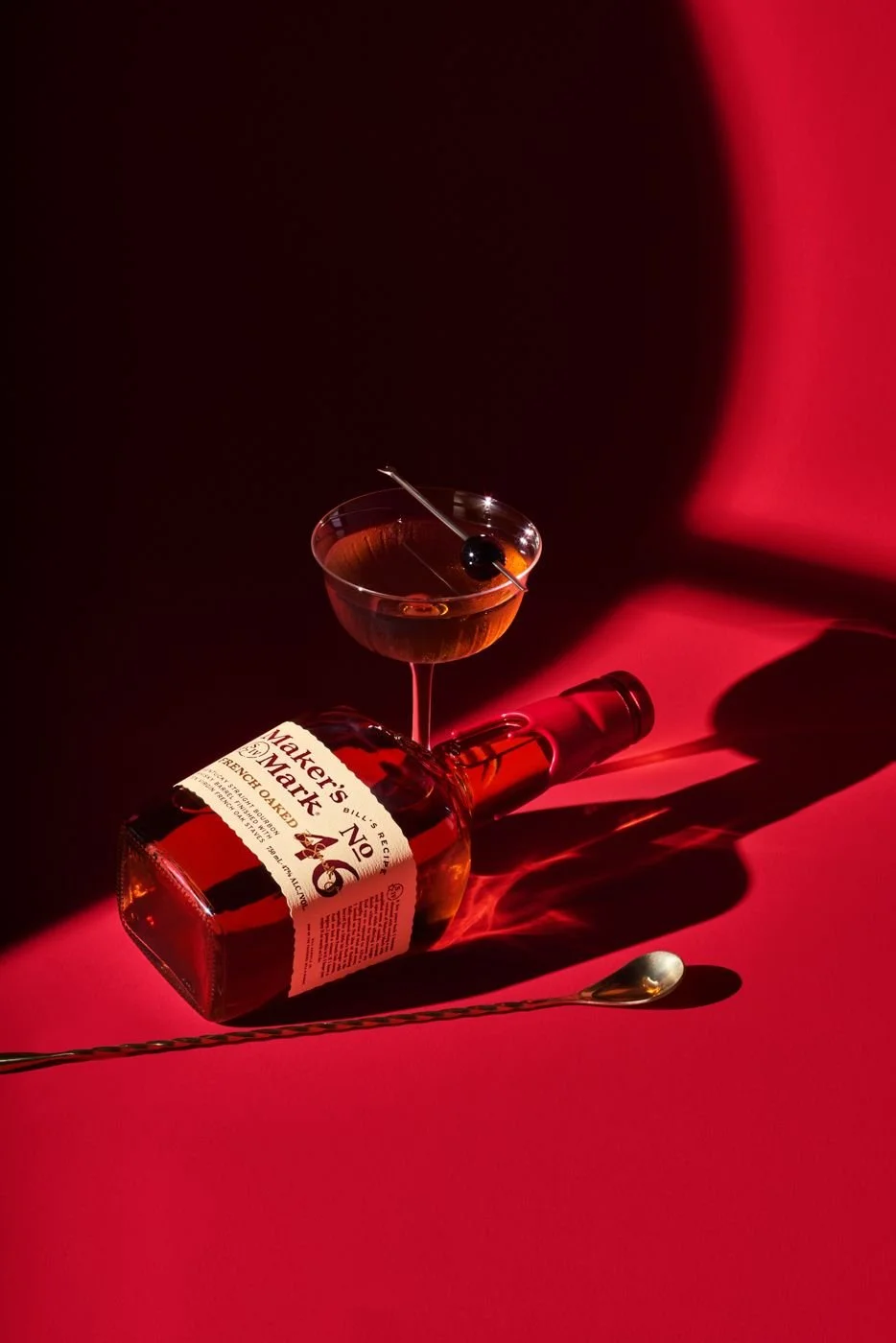 A bottle of Maker's Mark bourbon lying on its side on a red surface, with a glass of bourbon, a cocktail pick with a black cherry, and a long spoon nearby, all cast with dramatic shadows in a red and black background.