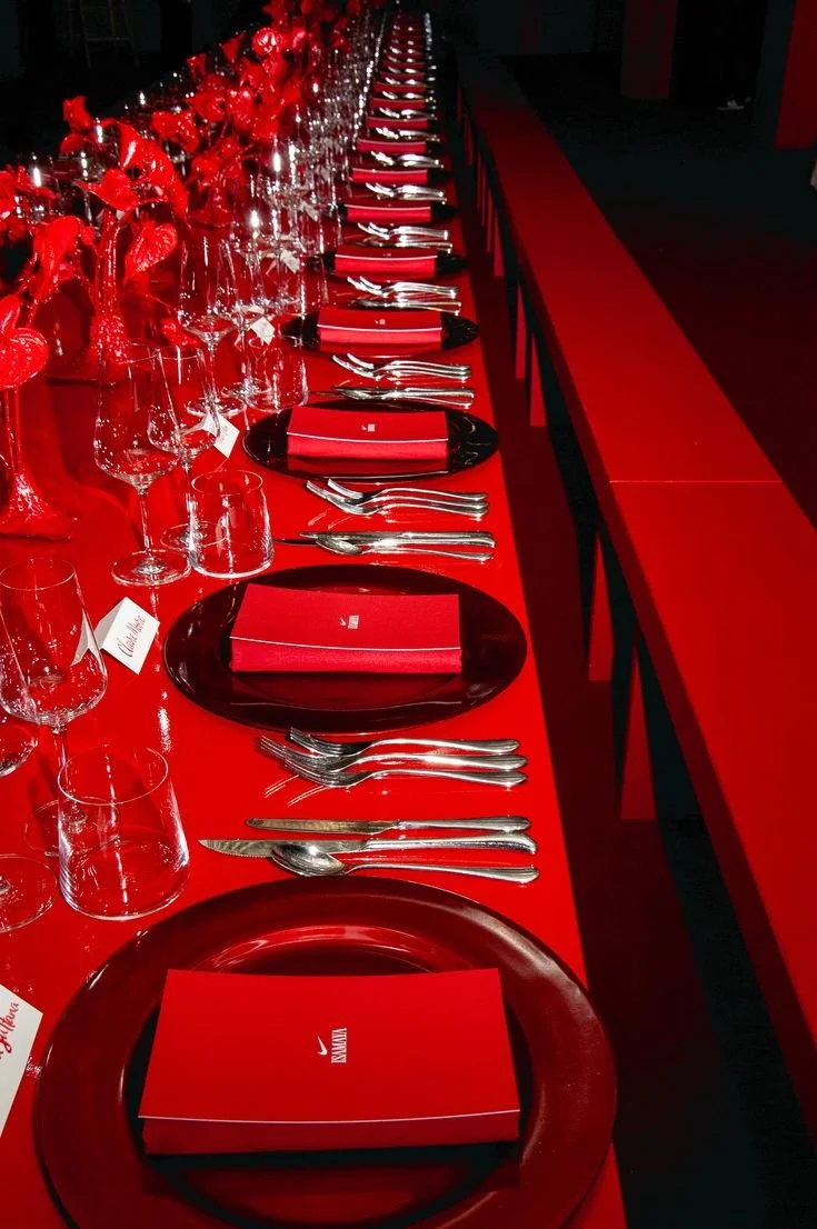 A long banquet table set for a formal event, with black plates, red napkins, silverware, and wine glasses. The table is decorated with red floral centerpieces.