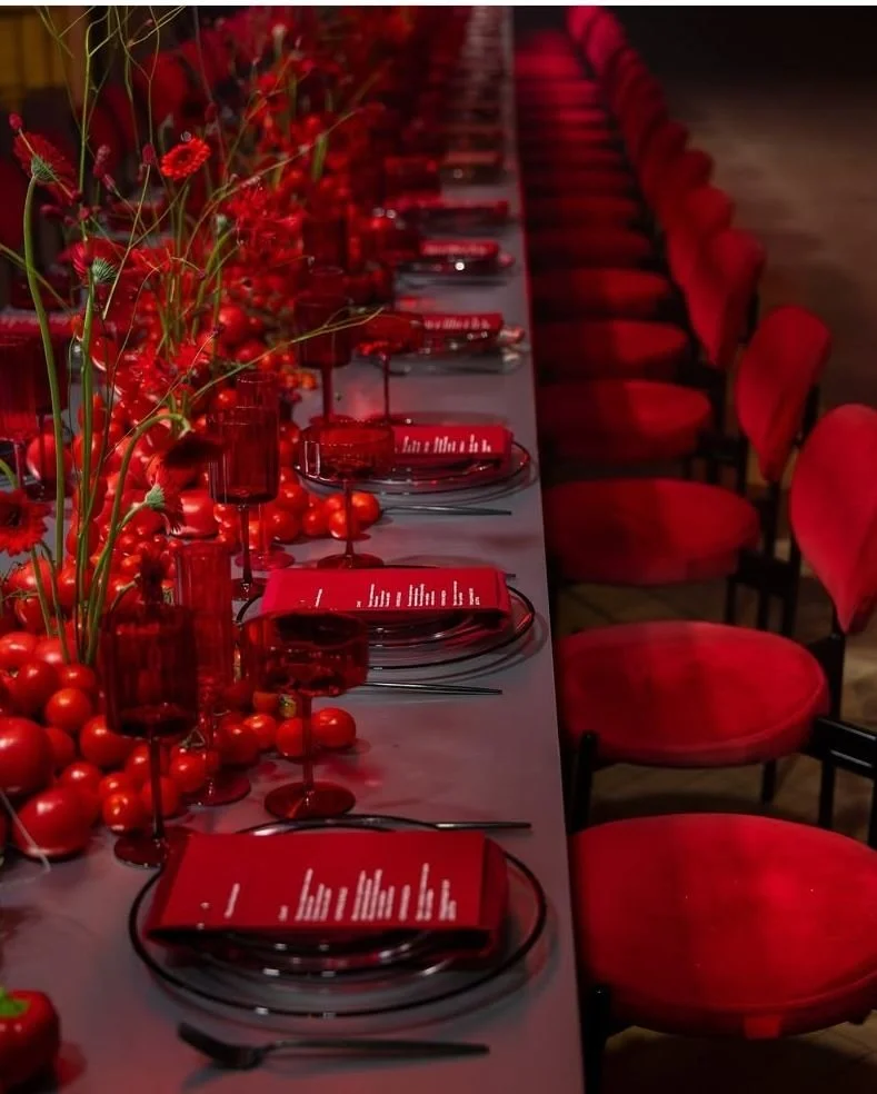Long dining table set for a formal event with red plates, glasses, and napkins, decorated with red flowers and fruit-based centerpieces, surrounded by red chairs.