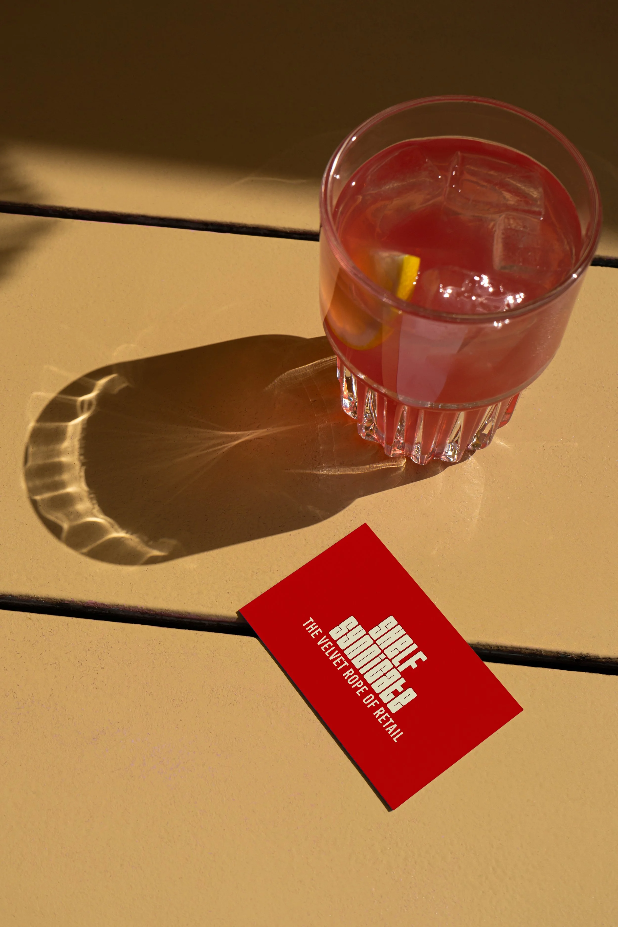 A glass of pink cocktail with ice cubes and lemon wedge casting a patterned shadow on a beige surface. A red card with white text reads 'SHIFT SINGO SU SING THE VELVET ROPE OF RETAIL' placed on the surface.