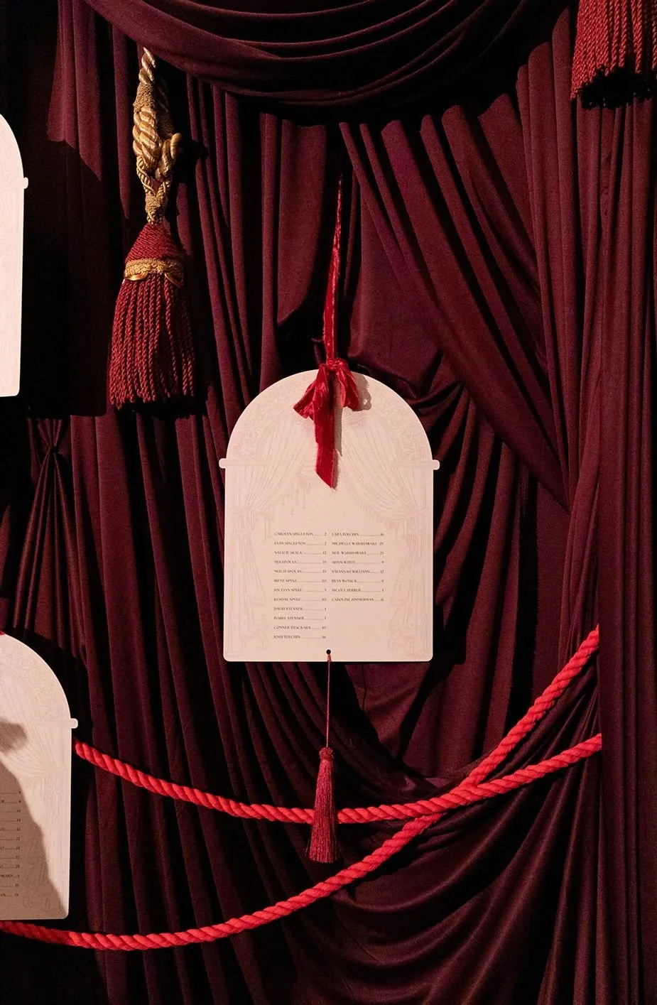A program or schedule hanging in front of dark red velvet stage curtains, decorated with a red tassel and rope, with some text and a large red ribbon at the top.