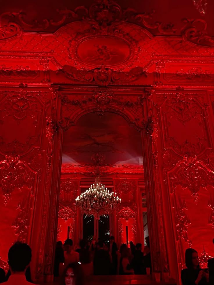 A lavish room with ornate wall and ceiling decorations illuminated in red light, chandelier hanging from the ceiling, and people gathered inside.