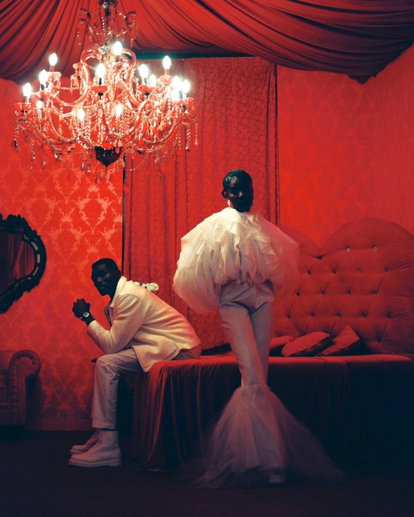A woman in a dramatic white gown standing on a bed in a richly decorated room with red wallpaper and curtains, illuminated by a large chandelier, while a man in a white suit sits on the edge of the bed.