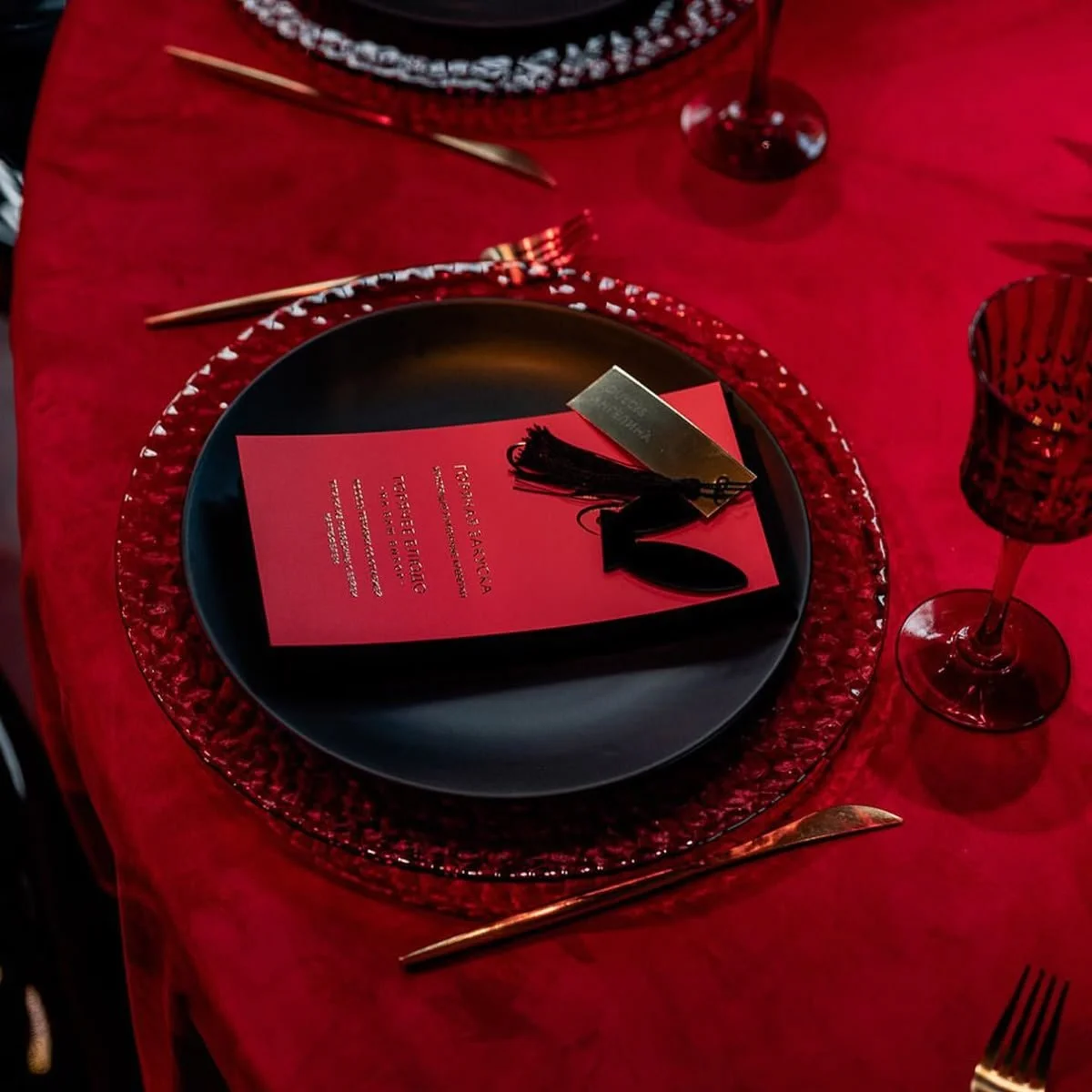 A formal table setting on a red tablecloth with a black plate, decorative red glass plate, gold utensils, red wine glass, and a red program or menu with black tassel on top.