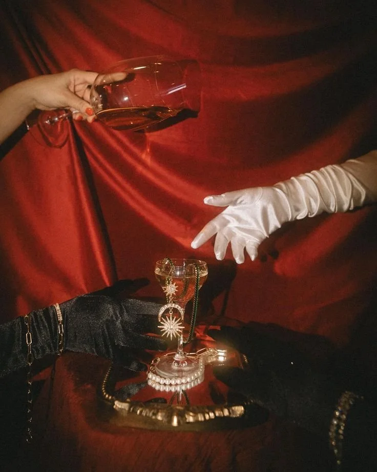 Two hands, one with orange nail polish, are reaching for a crystal glass of champagne on a dark wooden table. One hand wears a long white glove, and the other a black glove with jewelry. The background is red velvet fabric.