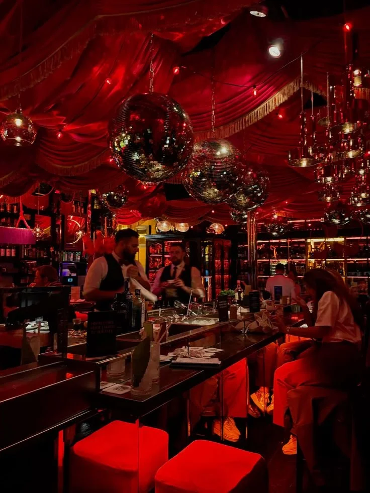 Interior of a dimly lit bar or lounge with red lighting, reflective disco balls hanging from the red draped ceiling, and patrons sitting at the bar.