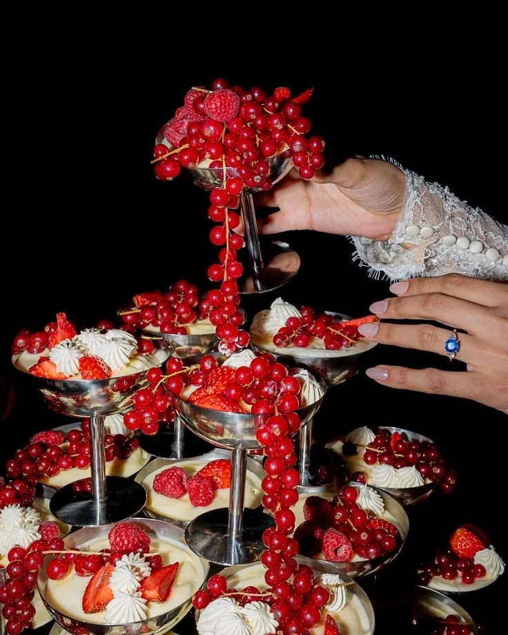 A hand holding a silver bowl filled with red berries, pouring onto a display of many other bowls filled with strawberries, raspberries, whipped cream, and red berries against a black background.