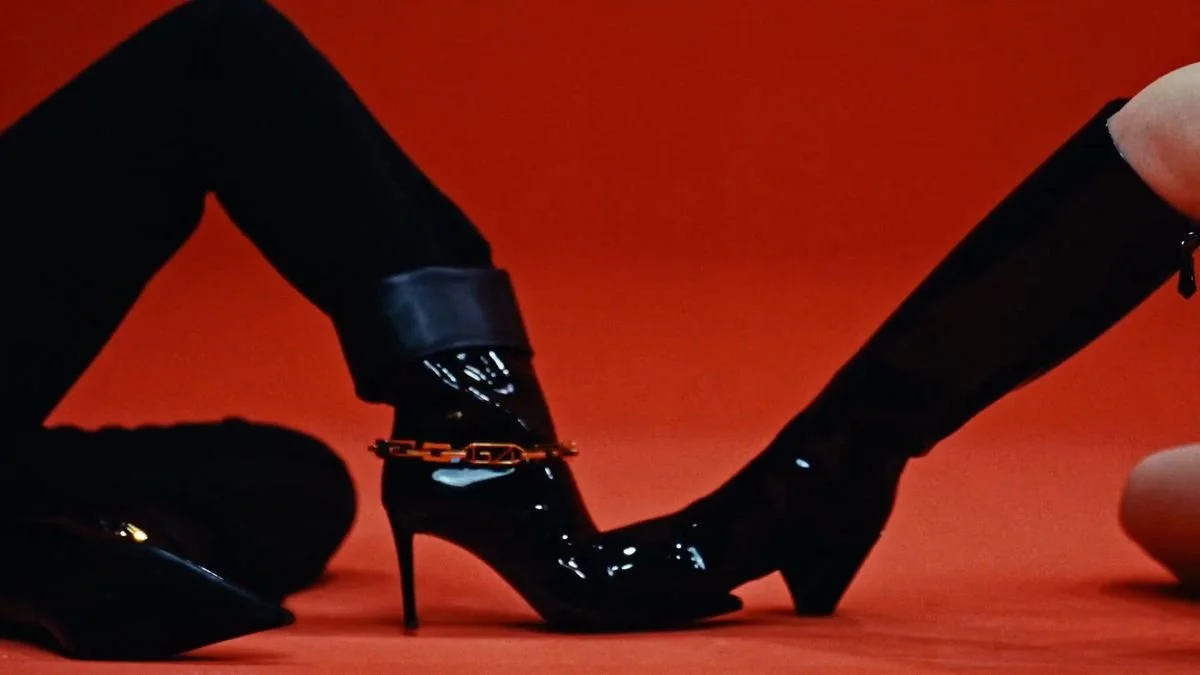 Close-up of a person wearing black high-heeled boots and black pants, with gold chains around the ankle, against a red background.