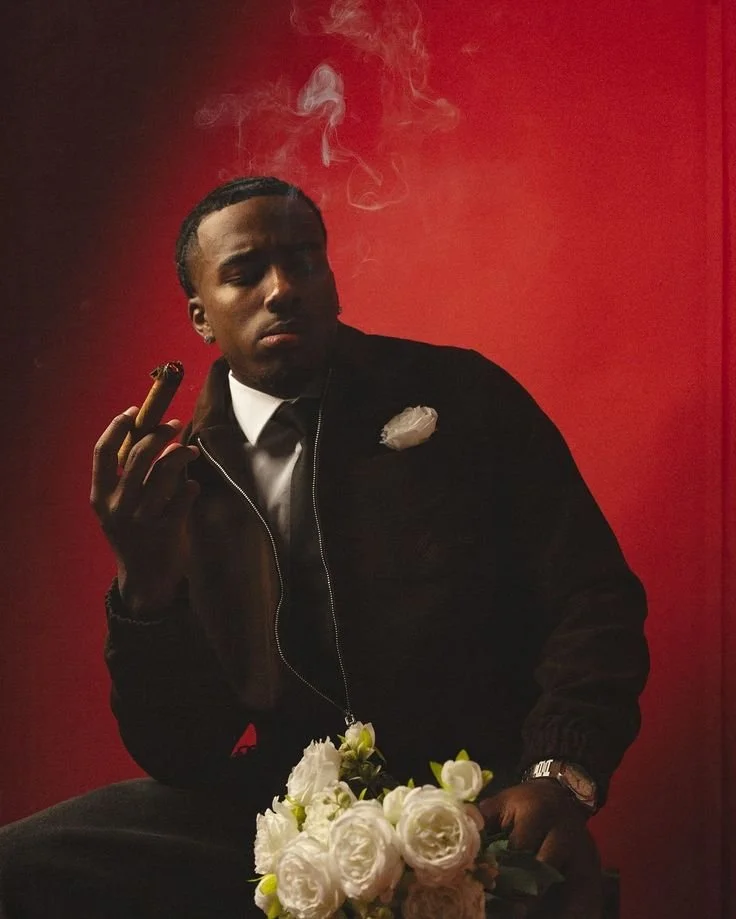 A young man with braided hair, wearing a black jacket and white shirt, sitting against a red background, holding a lit cigar in one hand, with a clutch of white flowers on his lap, and a white flower on his chest.