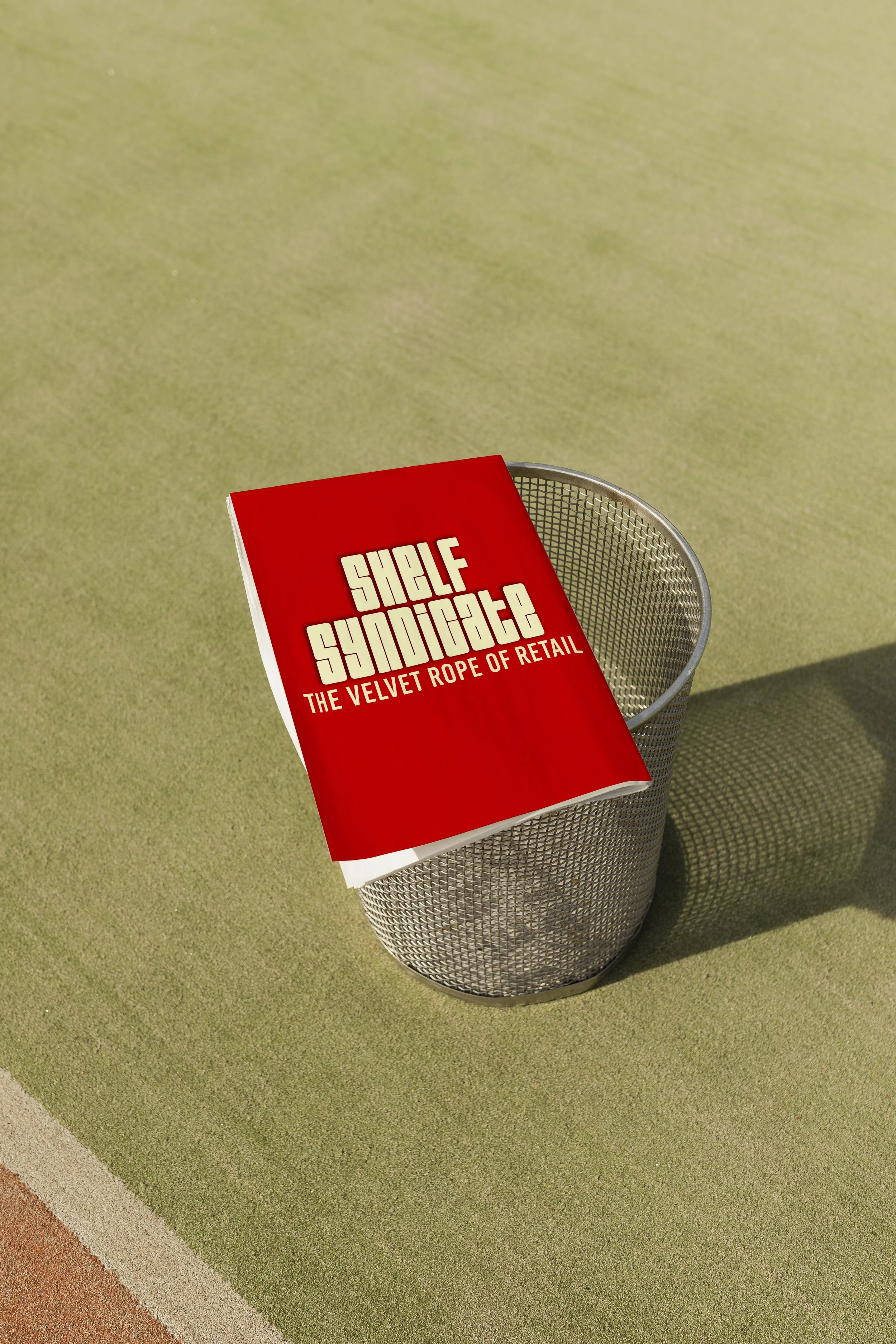 Red magazine titled "Shelf Syndicate: The Velvet Rope of Retail" resting on a metal mesh trash can on a green and tan floor.