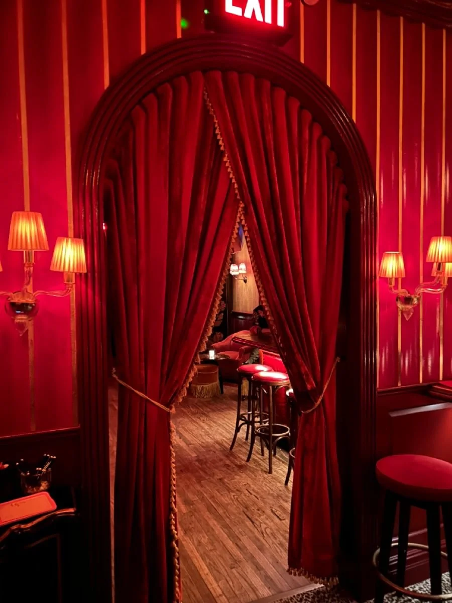 Red velvet curtain opening to a cozy bar with wooden stools and warm lighting, red walls with gold trim, and wall sconces with lampshades.
