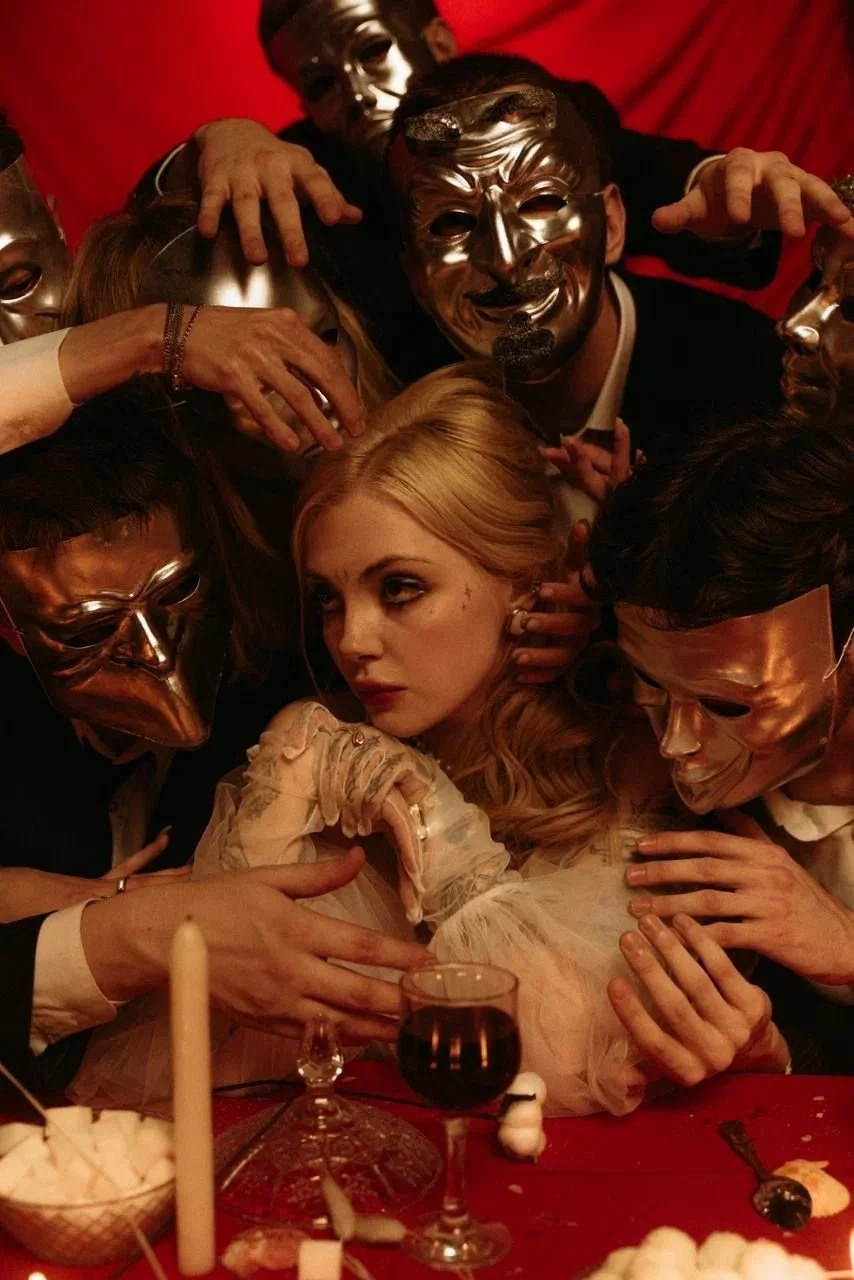 Group of people wearing metallic masks surrounding a woman with blonde hair, at a dimly lit event with red background and table with wine and snacks.