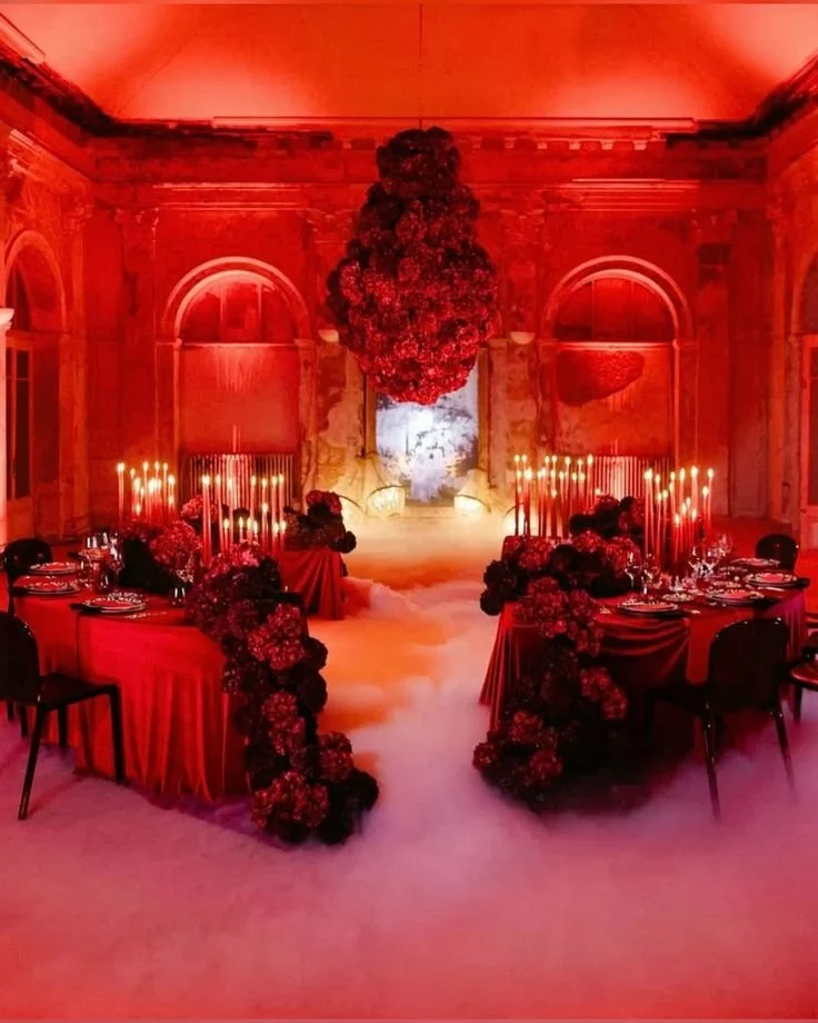 Elegant dining room with red lighting, decorated with floral arrangements, candles, and draped tablecloths, with a pathway in the center covered in white fog.
