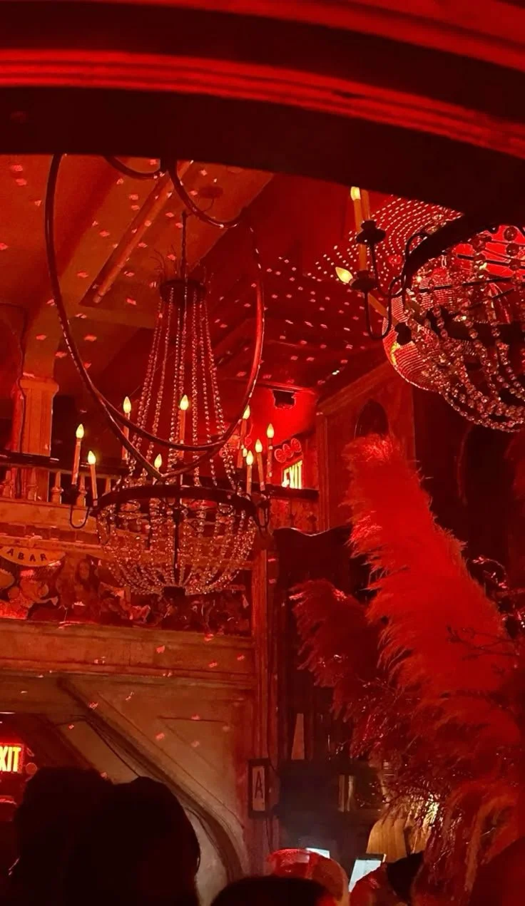 Chandeliers hanging from a ceiling, illuminated with warm yellow lights in a dimly lit, red-toned interior with people sitting and a decorative feather arrangement on the right side.