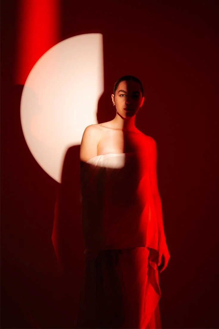 A woman standing in front of a dark red background, illuminated by a white crescent-shaped light source casting shadows on her face and body.