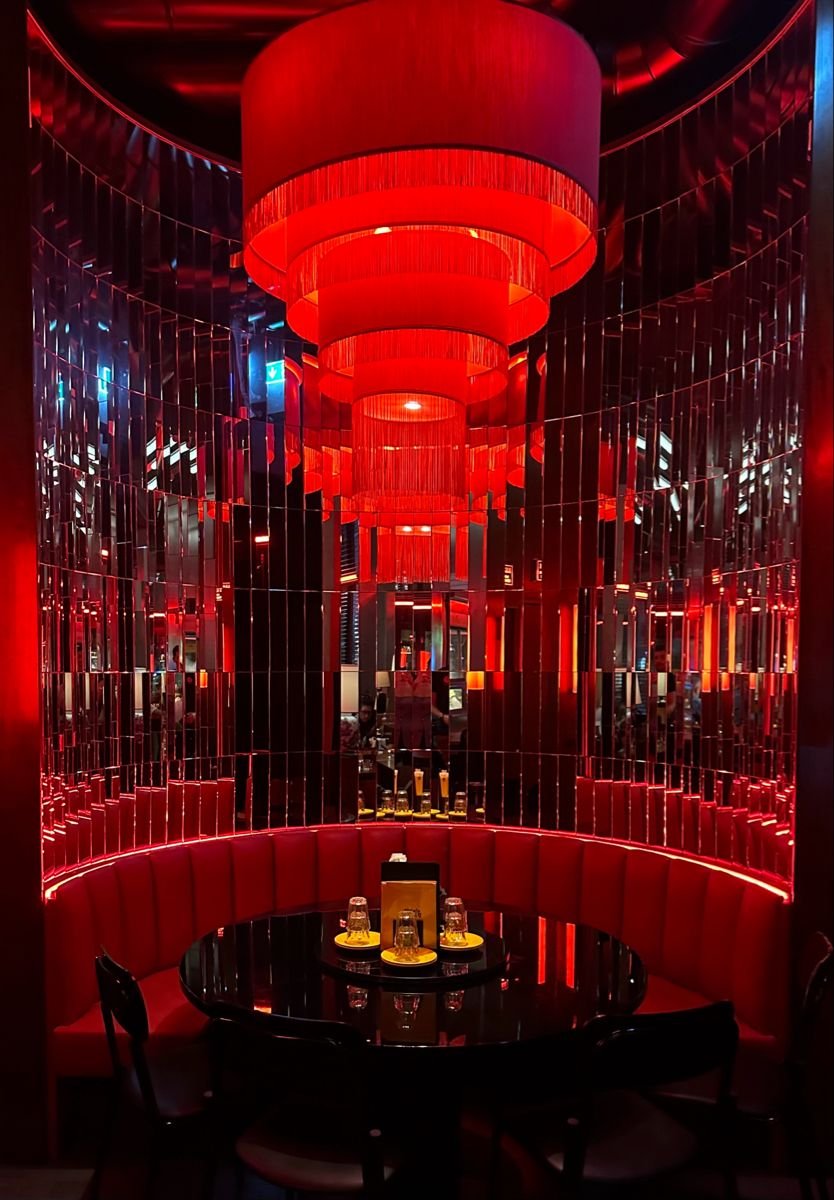 A luxurious restaurant corner with a round table set with glasses and napkins, surrounded by a curved red upholstered banquette. The decor is dramatic with red lighting, including large red drum-shaped hanging lamps and mirror panels creating a refle