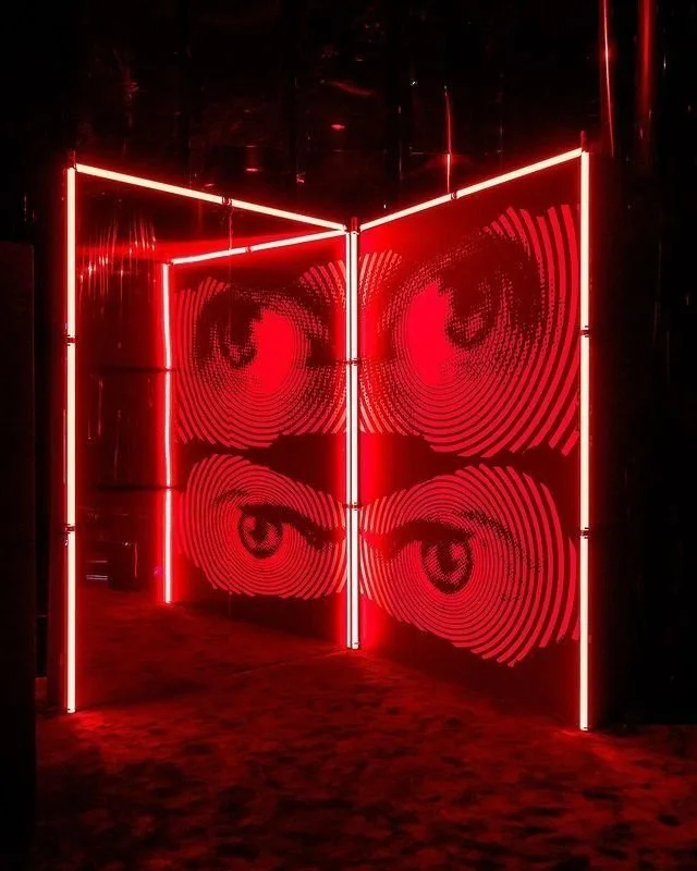 Light installation with four large red eyes created with concentric lines, arranged on a dark wall with vertical neon light supports.
