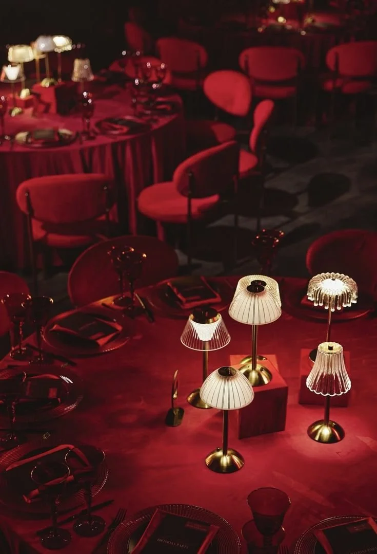 Elegant restaurant table setting with red chairs, dark tablecloths, and multiple small lamps providing warm lighting.