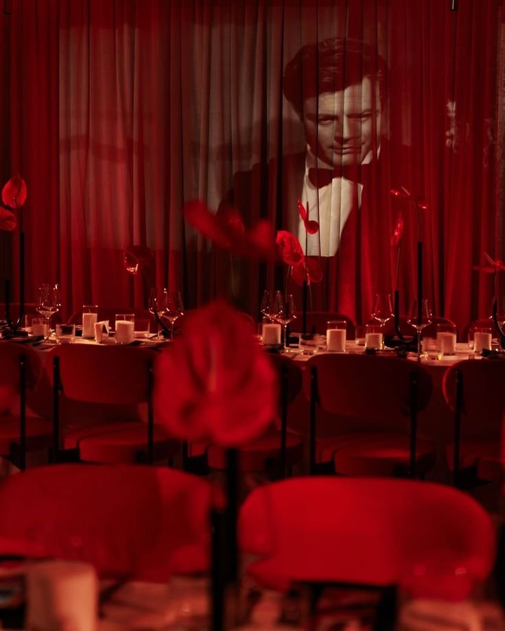 A dimly lit restaurant decorated for a romantic occasion, with a large black-and-white photo of a man in a tuxedo projected onto curtains behind the dining area, and red roses on the tables.