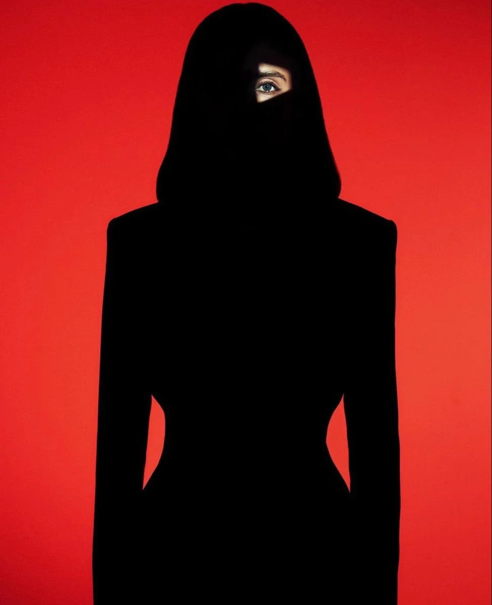 A woman wearing a black niqab with only one eye visible, standing against a red background.