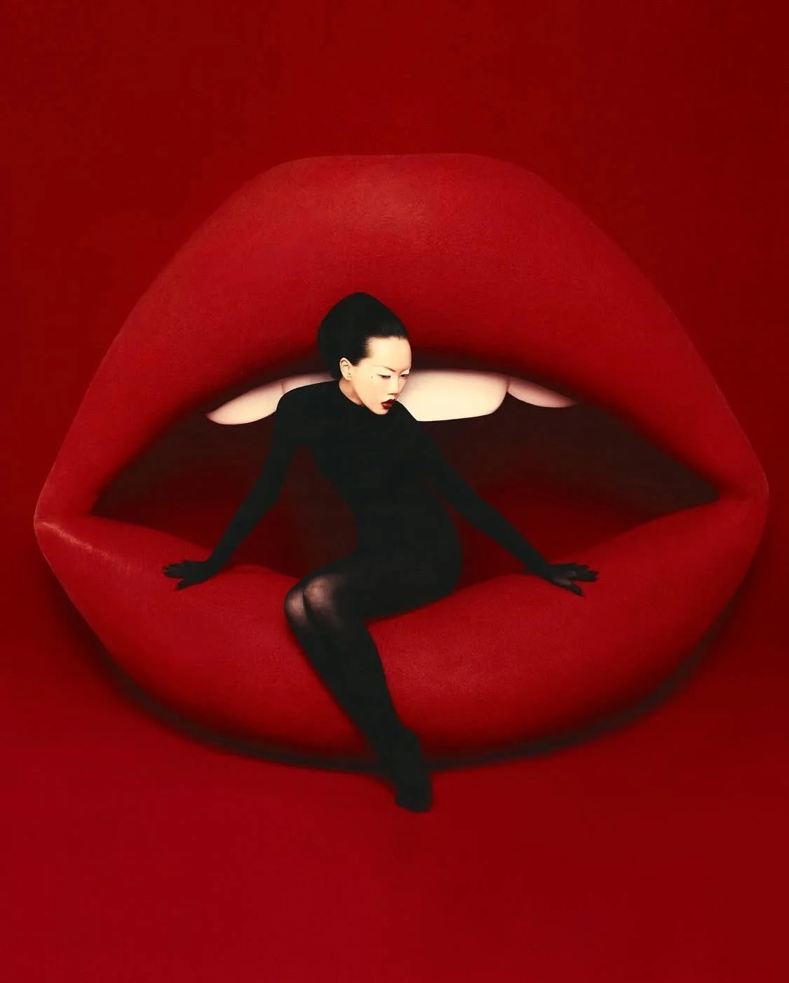 A woman dressed in black sitting inside large red lips with her arms outstretched.