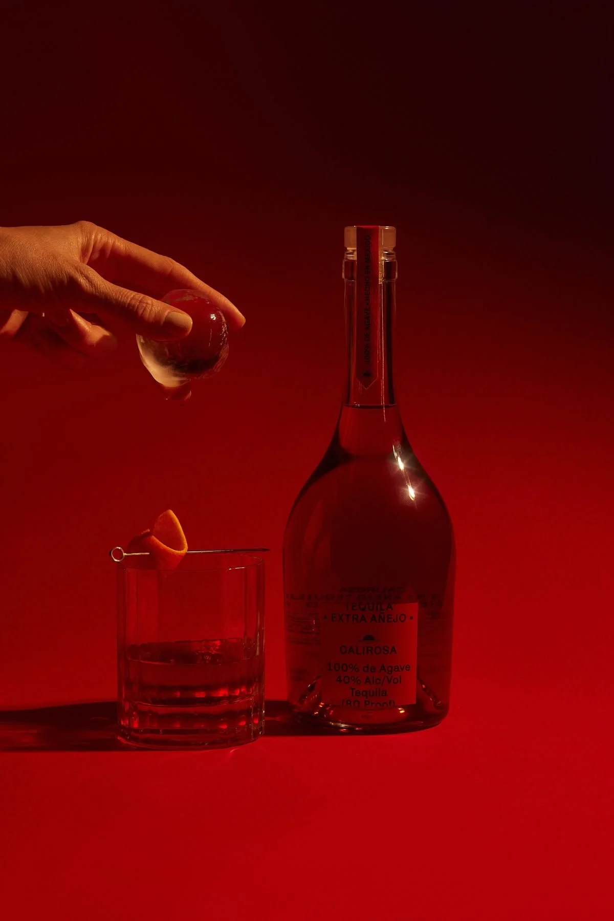 A hand placing a ball carved from a red fruit or vegetable into a glass of clear liquid, with a bottle of tequila and an orange slice garnishing the glass, against a red background.