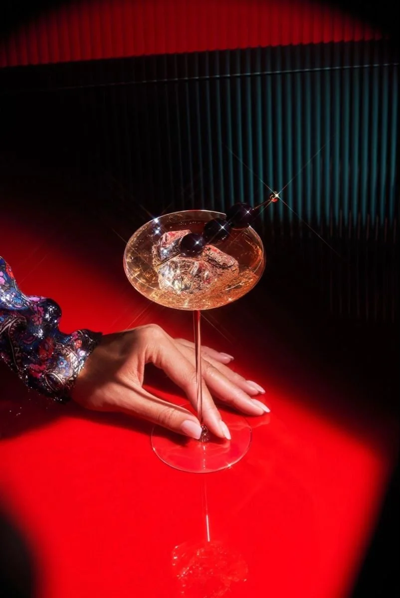 A person with a patterned sleeve holding a fancy cocktail glass with ice and three black olives, against a red and black background.