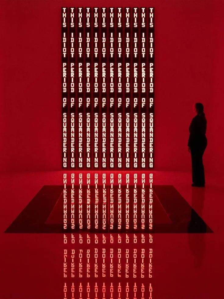 Text art installation on a red wall featuring the phrase "This I Do Not Perish" repeated in vertical columns, with a person standing to the right observing it.