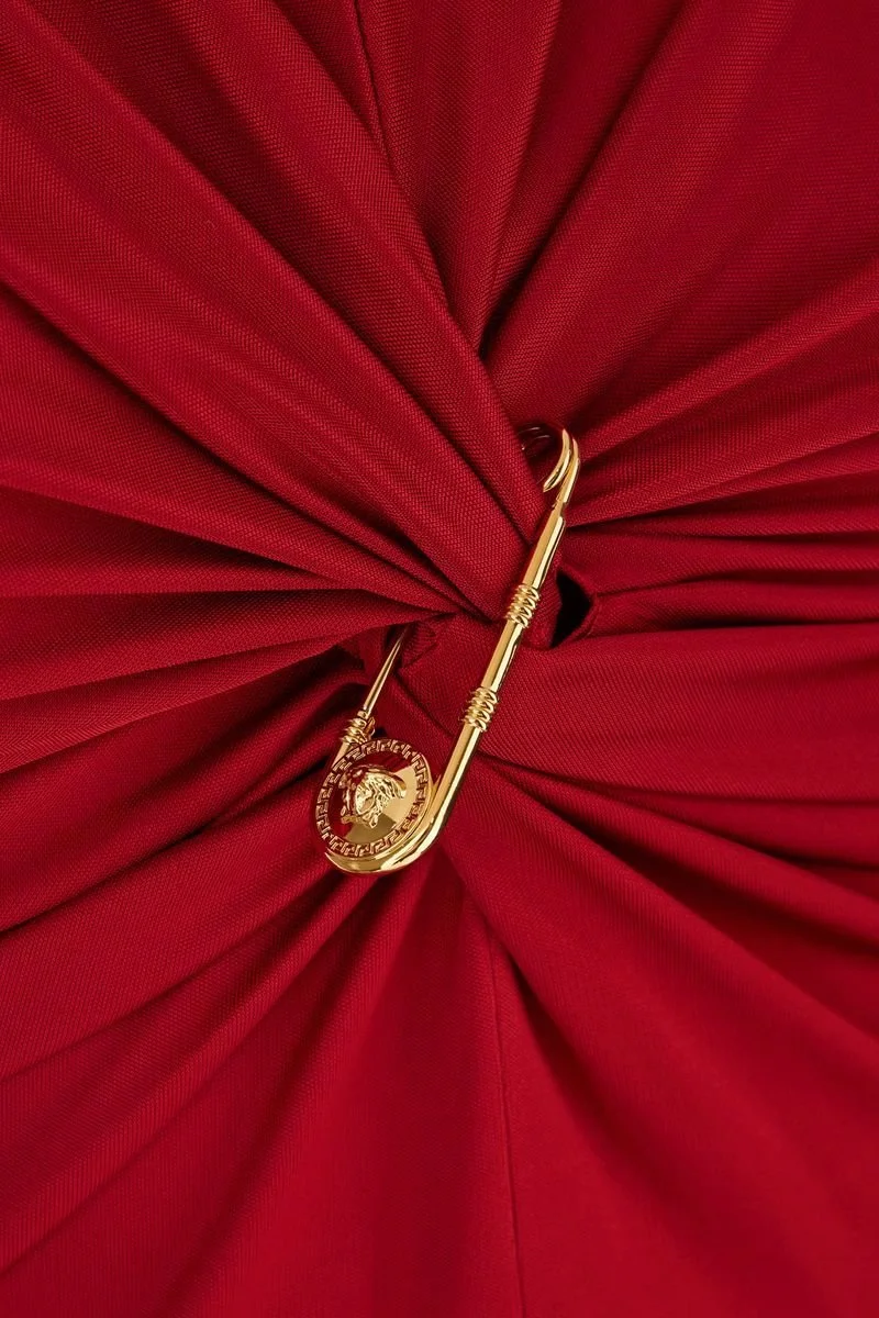 A gold safety pin with a Medusa head Medusa coin attached, pinned to a red fabric background.
