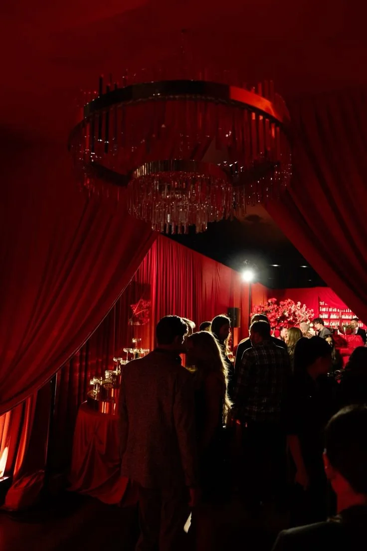 People mingling at an elegant party or event with red curtains, a chandelier, and floral decor.