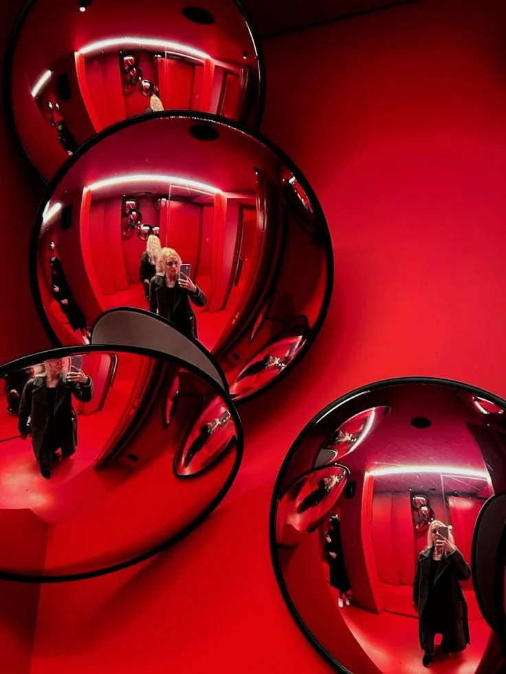 Reflection of a woman taking a selfie in multiple round mirrors on a red wall.