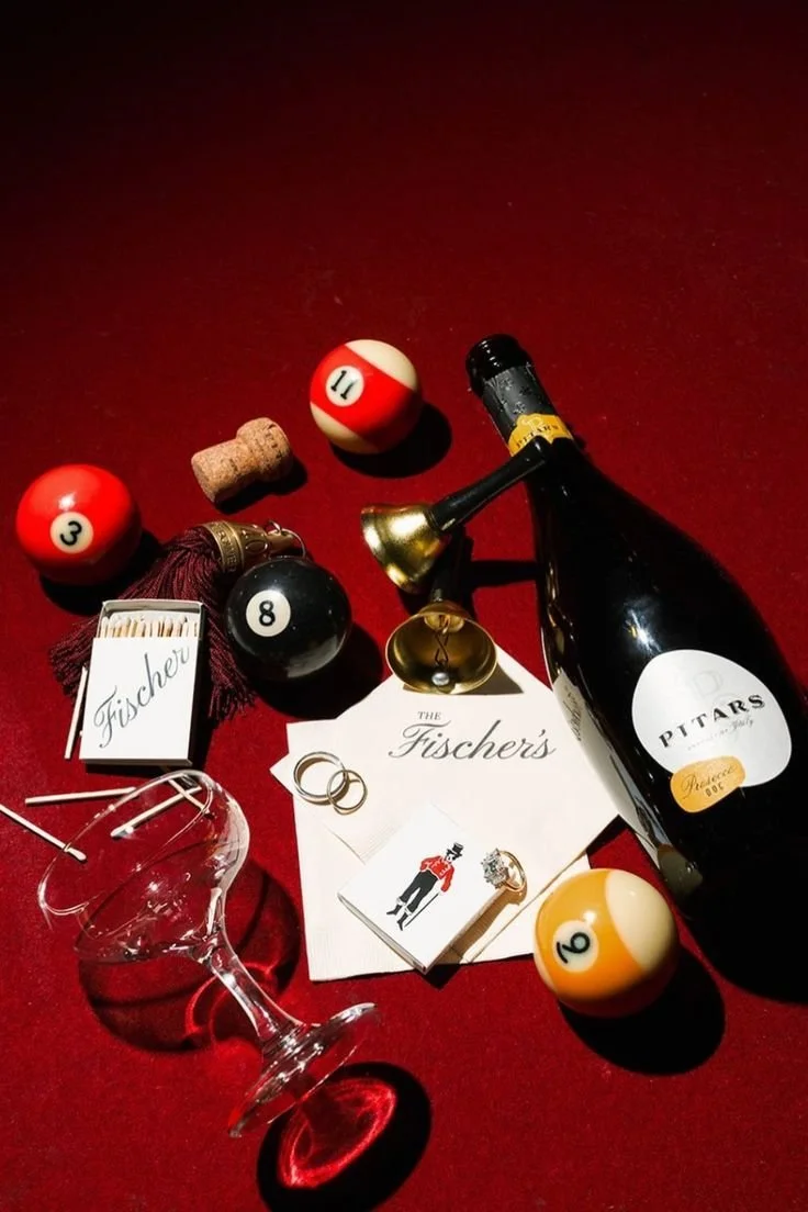 A bottle of wine, a glass, coasters, billiard balls, a cork, matches, and a small bell on a red surface.