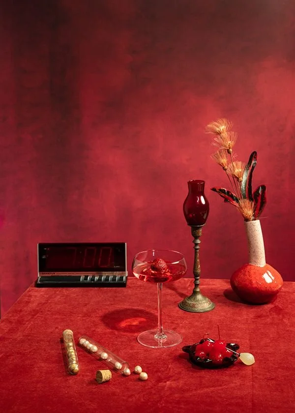 Red tablecloth with decorative items including a digital clock showing 7:00, a cocktail glass with a red drink, a tall red candle holder, a large red vase with dried plants, a glass jar with pearls, a cork, a red and black ornamental dish with small 