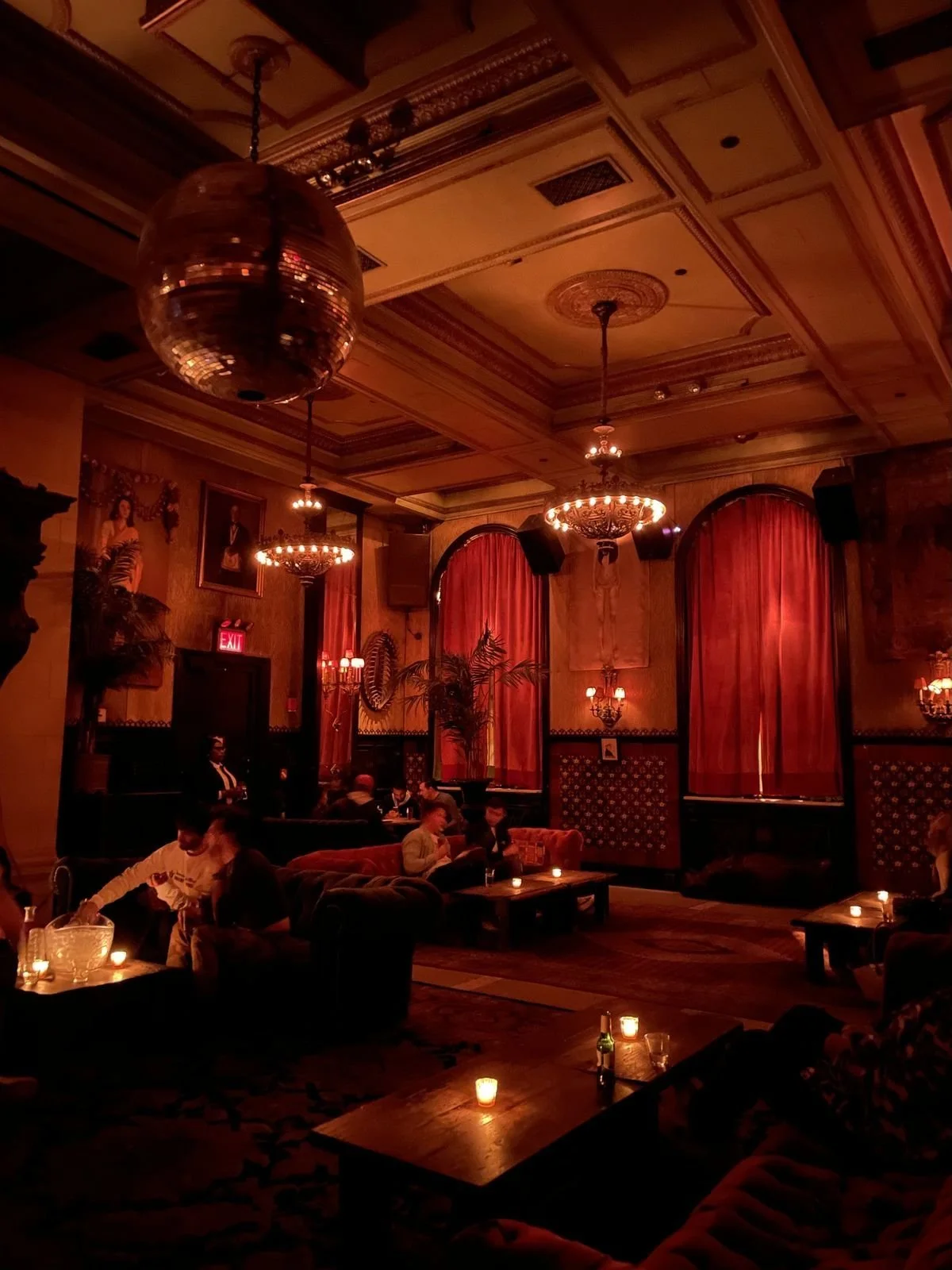Interior of a dimly lit lounge or bar with red curtains, chandeliers, and people sitting on sofas and tables, candles providing ambient lighting.