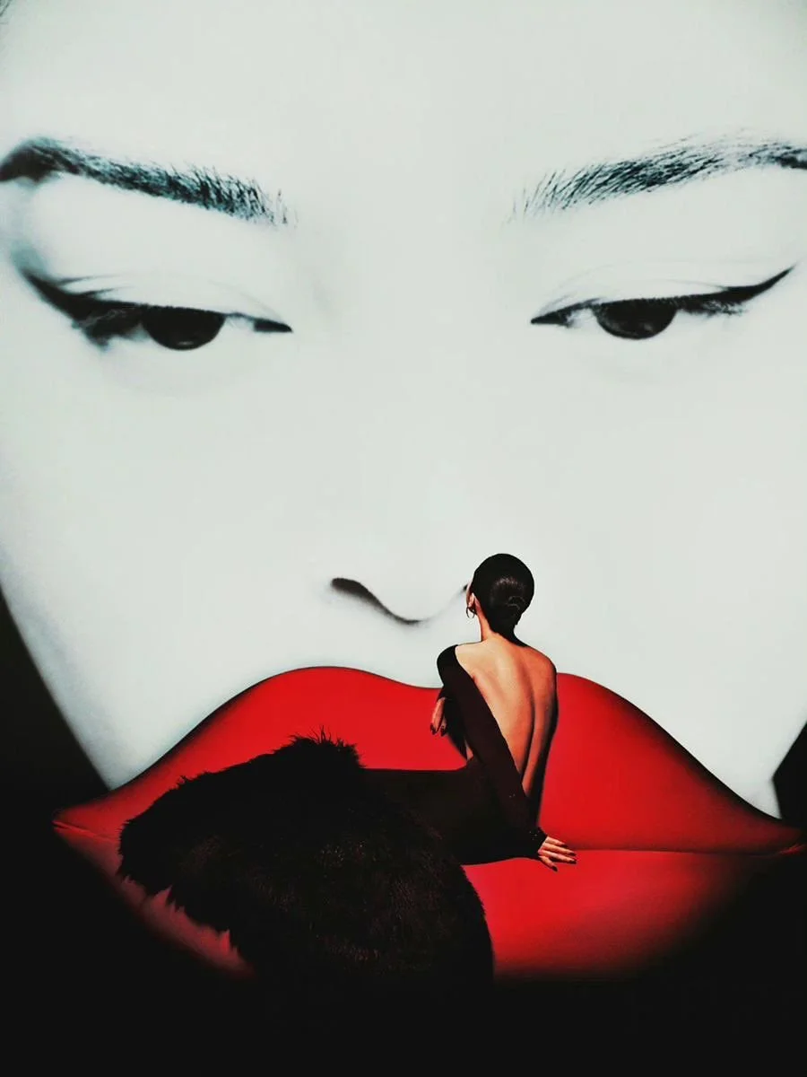 A woman with her back to the camera and short dark hair, sitting on a red surface, looking at a large black and white display of a woman's face with closed eyes and red lips.